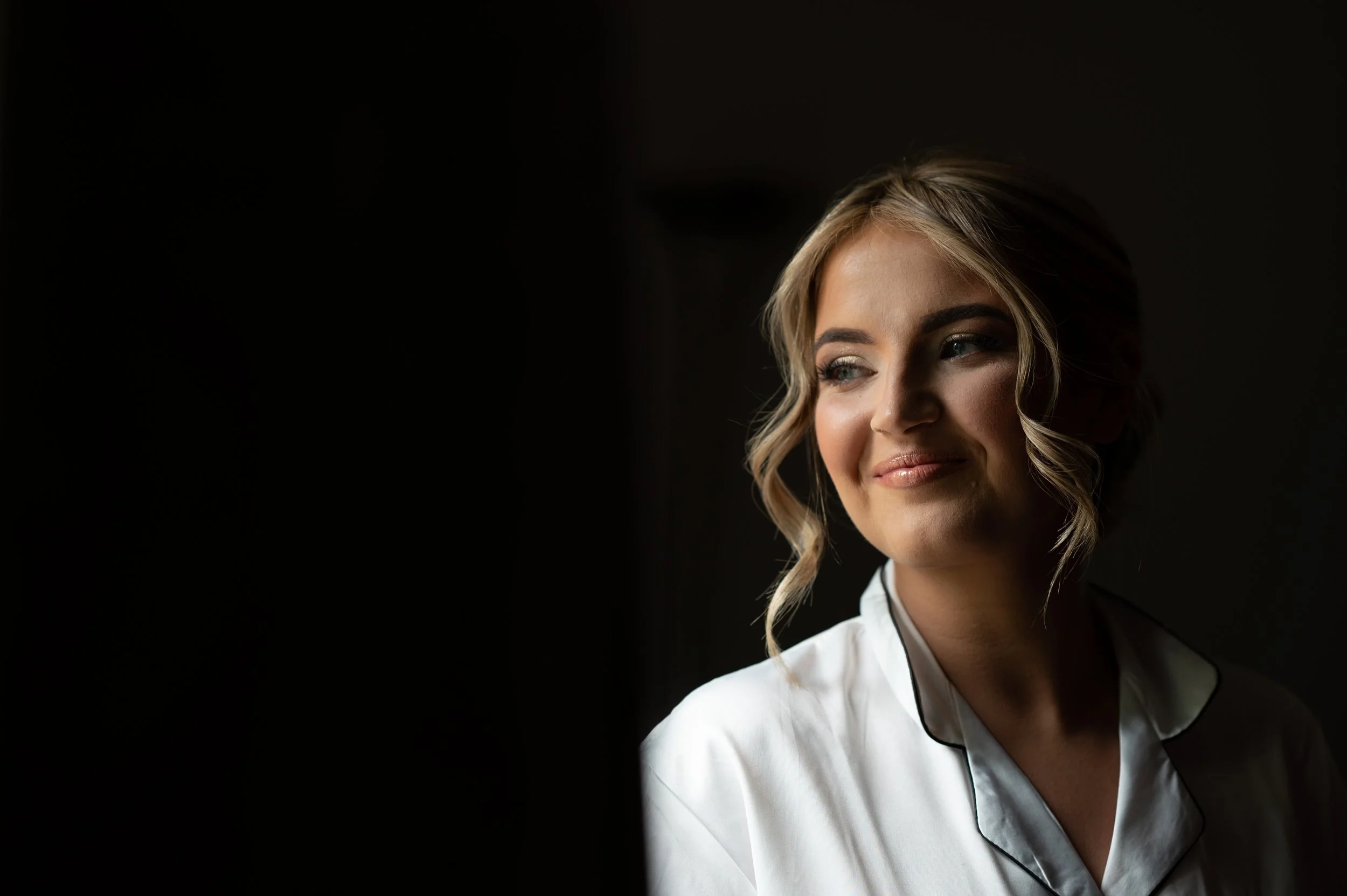 A woman with blonde, wavy hair styled in a side part, wearing a white blouse with a black piping detail, smiling softly, with a dark background.