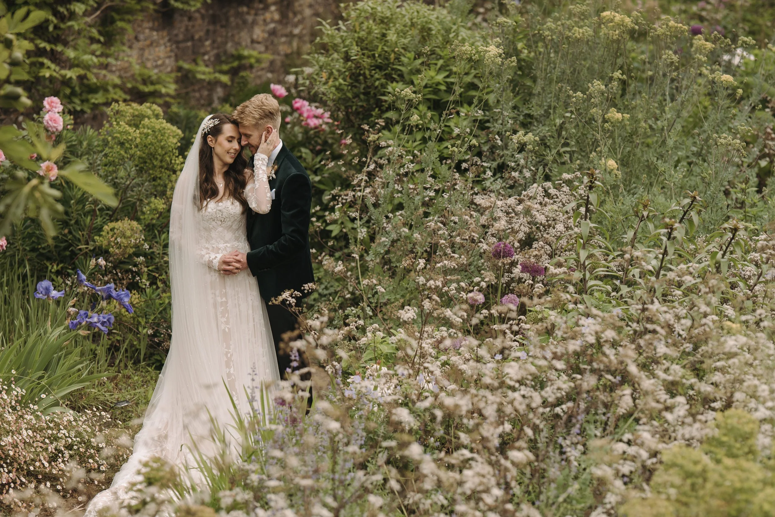 A bride and groom embrace under a canopy of flowers and greenery, with the bride in a white wedding dress and veil, and the groom in a black suit, touching foreheads in a garden setting.
