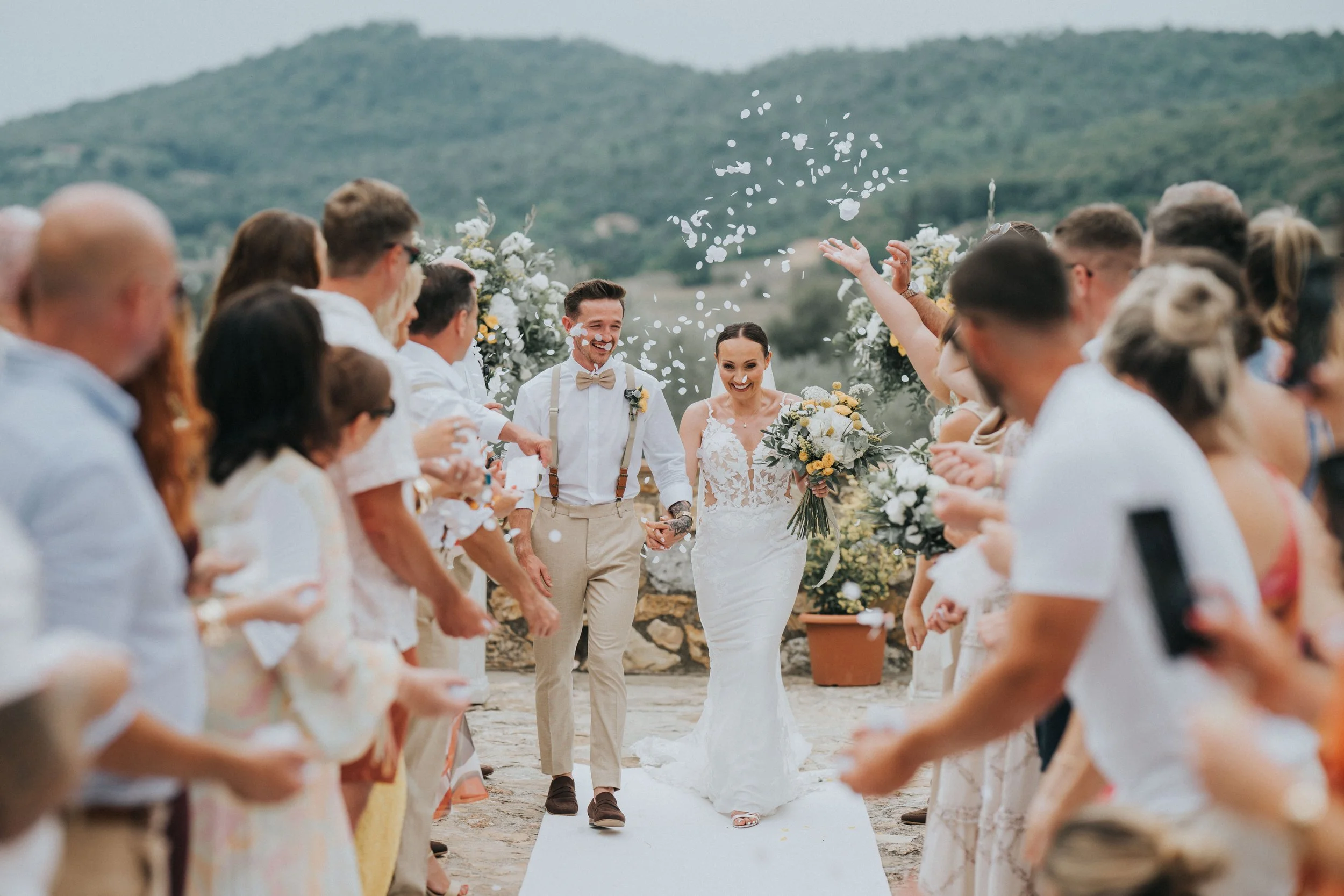 A bride and groom celebrating during their outdoor wedding ceremony with guests on either side, throwing flower petals and confetti. The groom is dressed in beige trousers, suspenders, and a bow tie, while the bride is in a white lace dress holding a