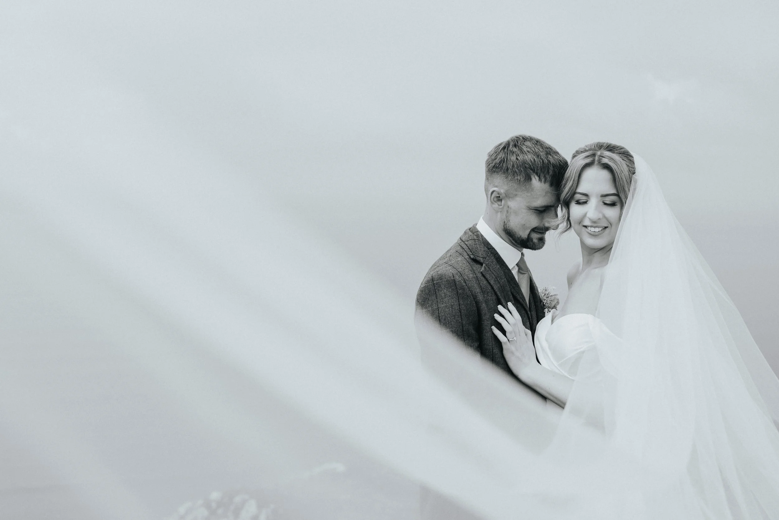 A black and white photograph of a bride and groom close together, with their foreheads touching and eyes closed, on a plain, light background.