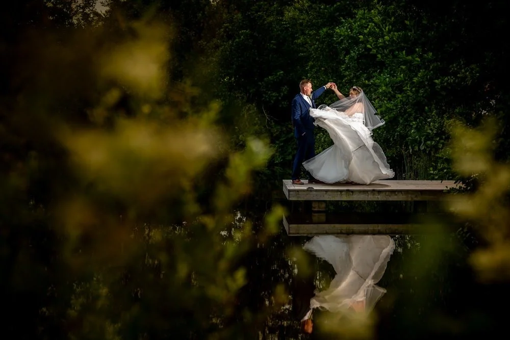 A bride and groom dancing on a small dock over a calm body of water, surrounded by green trees, with their reflection visible in the water.