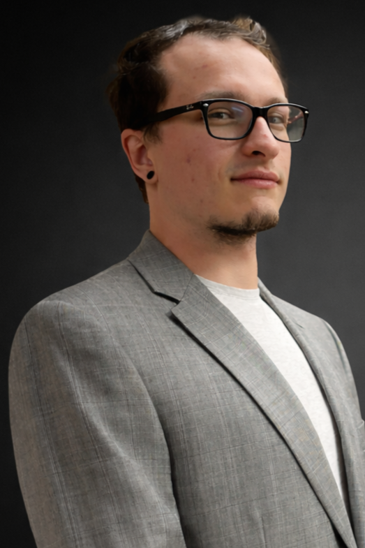 A young man with glasses and stretched earrings wearing a light gray blazer and a white t-shirt, standing against a dark background.