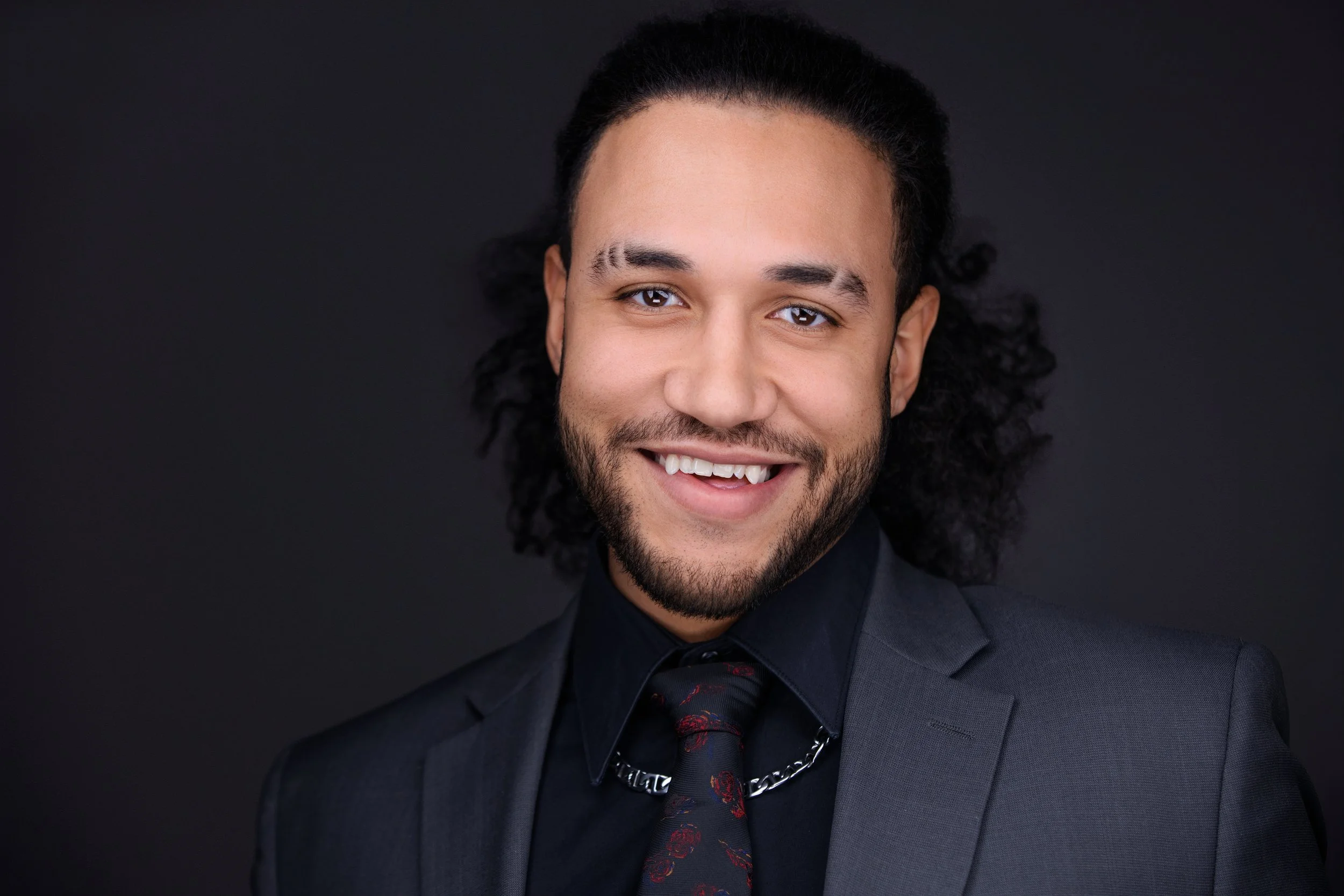 Portrait of a young man with long curly hair, wearing a black suit, a black shirt, a patterned black tie, and a silver chain, smiling against a dark background.