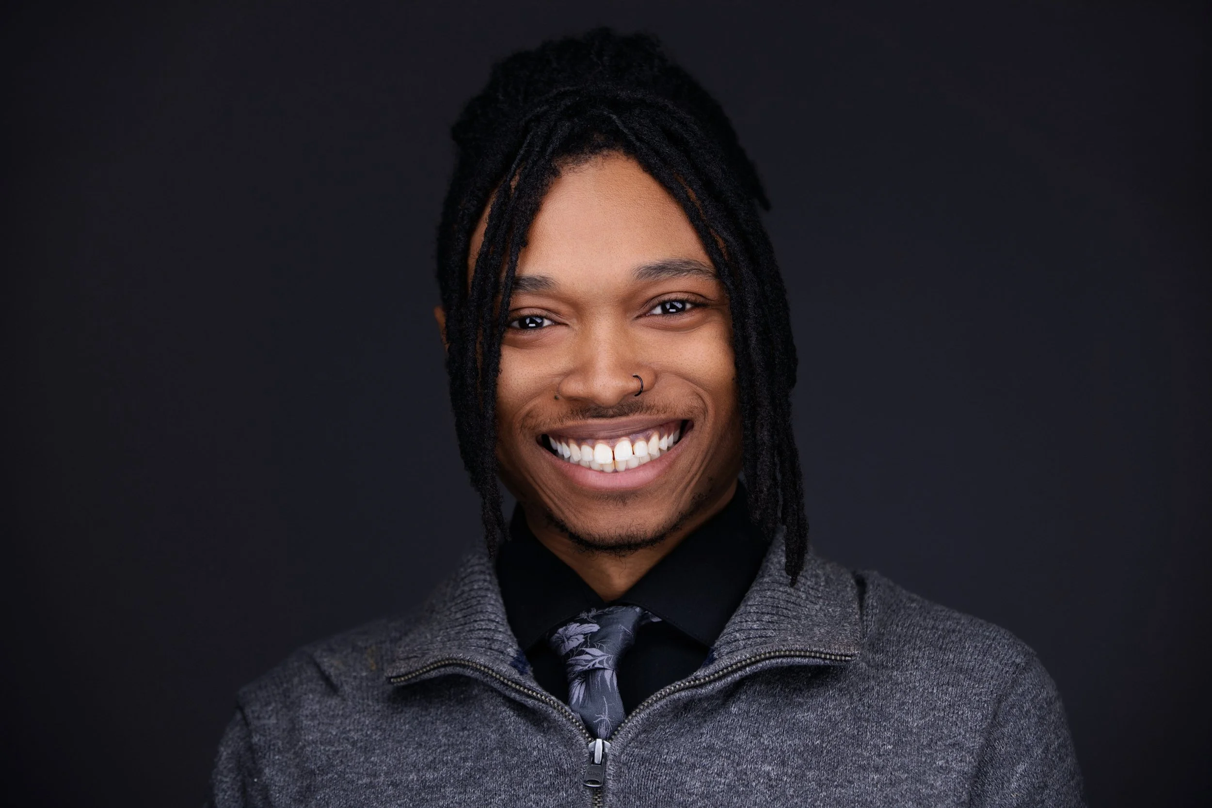 Smiling man with dreadlocks wearing a gray jacket, black shirt, and patterned tie against a black background.