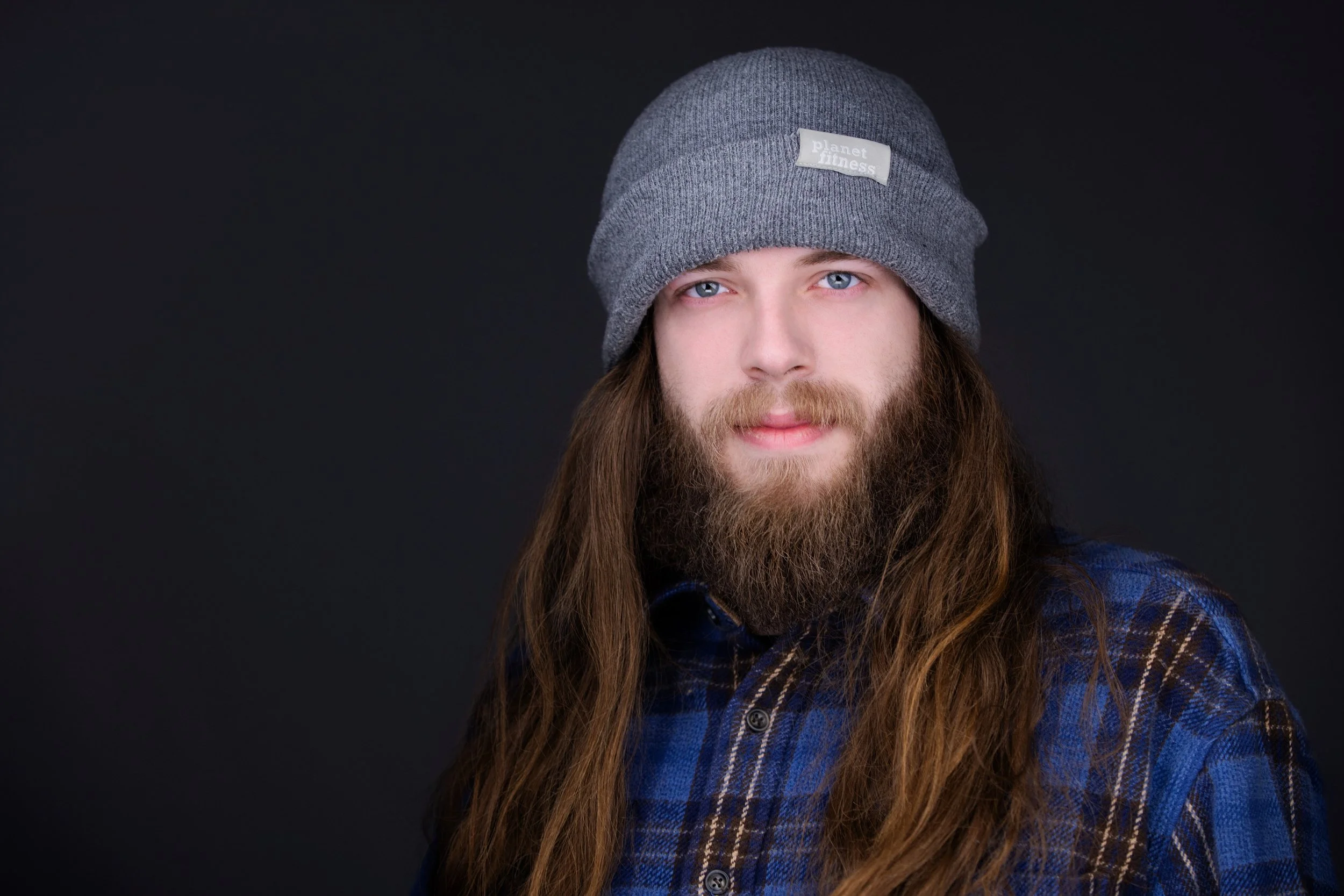 A young man with long brown hair and a beard, wearing a gray knit beanie and a blue plaid shirt, looks at the camera against a dark background.