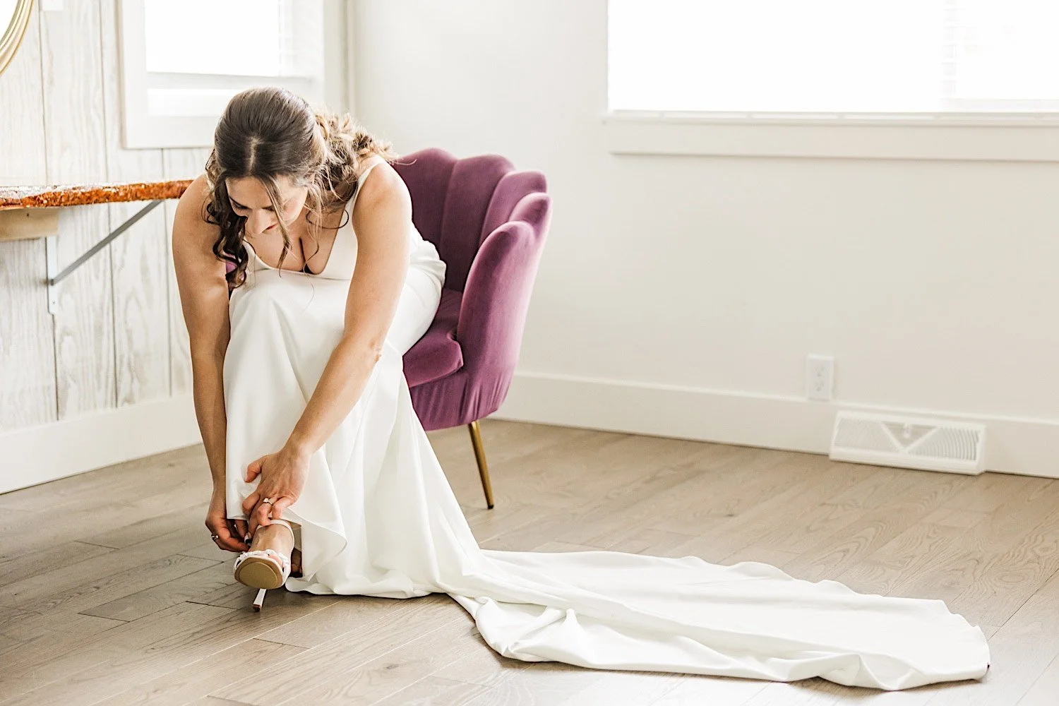 A bride sits in a purple chair to put on her wedding shoes