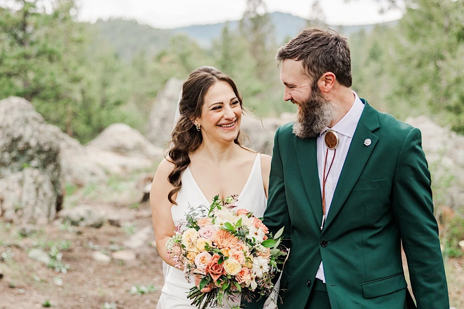 A bride and groom smile at one another while holding hands and walking along a trail in the mountains of Colorado
