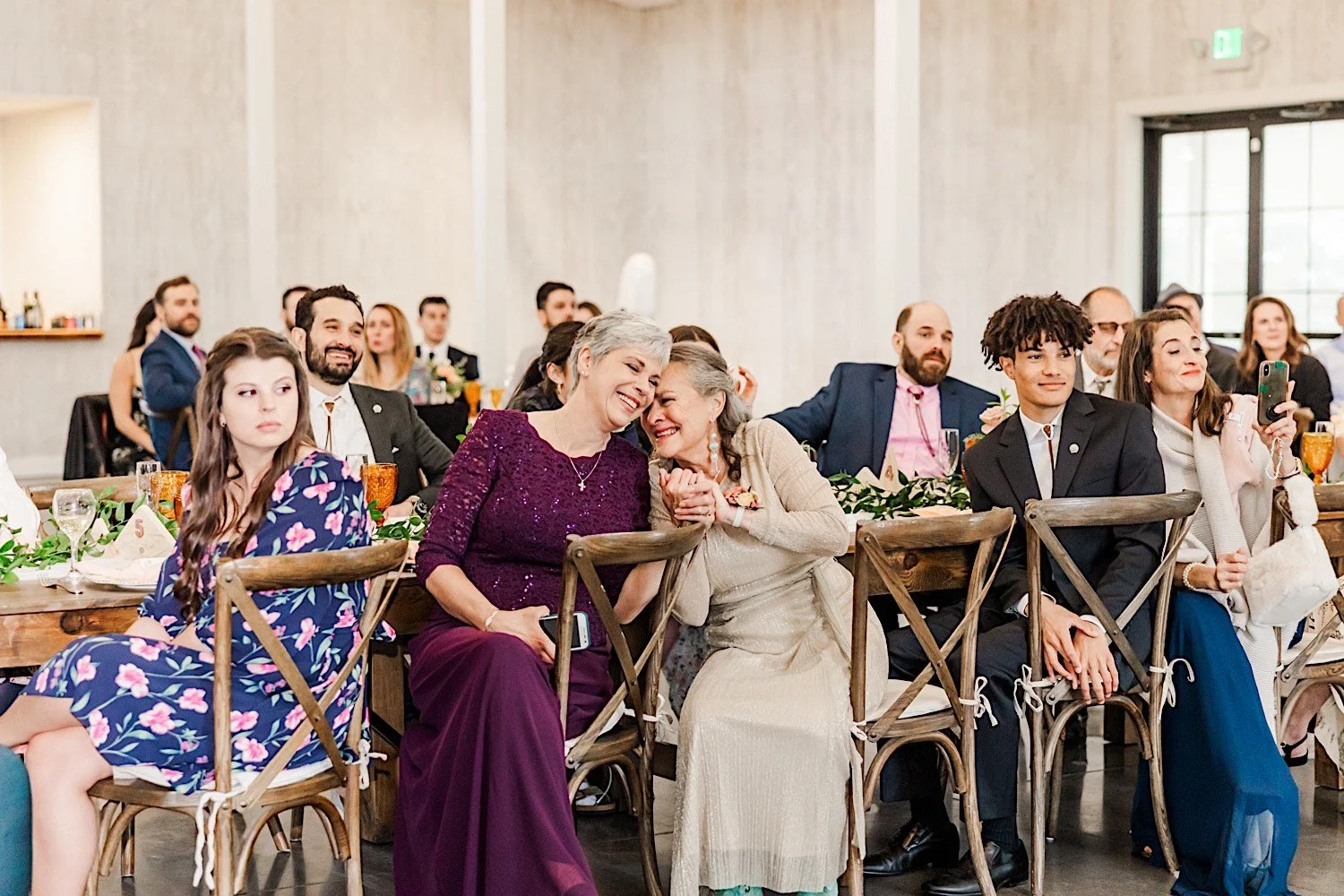 Mothers of the bride and groom sit next to one another to hold hands and smile as their children dance together during their indoor wedding reception at Woodlands Colorado