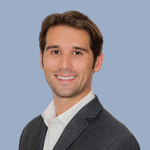 A smiling man with brown hair, wearing a dark gray suit and white shirt, standing against a light blue background.