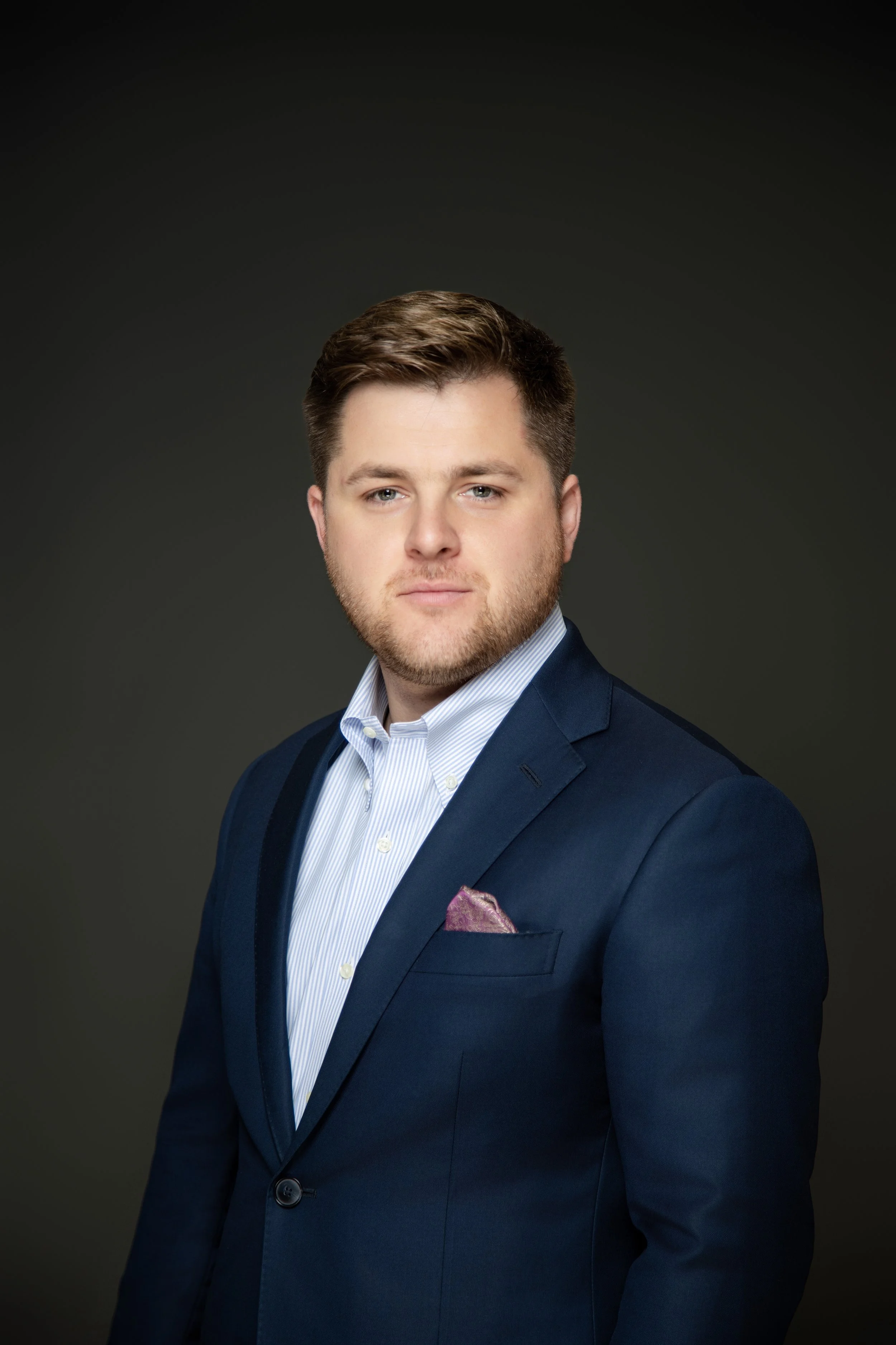 Headshot of a young man with brown hair and beard wearing a navy blue suit, light blue shirt, and a pink pocket square, against a dark background.