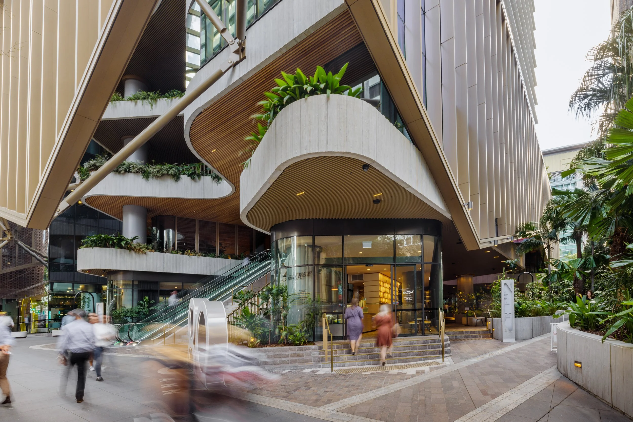 Modern, multi-story building with curved architecture, large glass entrance, and lush greenery, located in an urban environment with people walking nearby. Interior Designers in Brisbane Australia