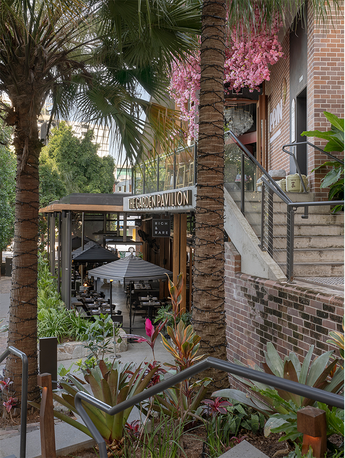 Outdoor restaurant seating under large black umbrellas, surrounded by lush green plants and palm trees, with a brick building in the background.