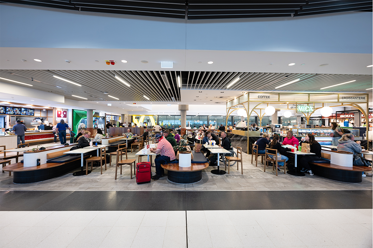 Interior of a busy airport food court with diners seated at tables and various food outlets visible, including coffee and croissant shops. Brisbane Airport Domestic Terminal. Retail Interior Design Australia