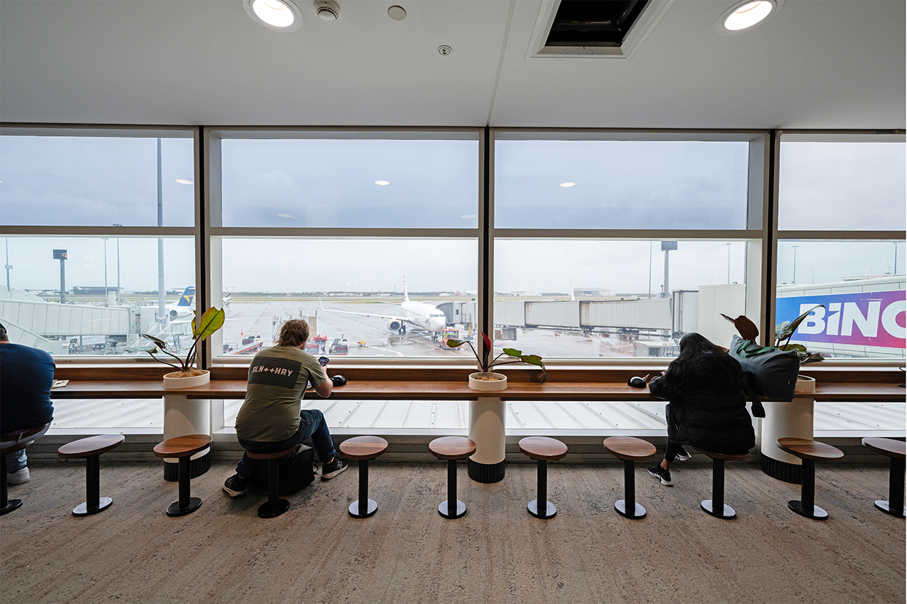 People seated in an airport lounge area, overlooking a runway with airplanes parked at the gate. Brisbane Airport Domestic Terminal. Retail Interior Design Australia