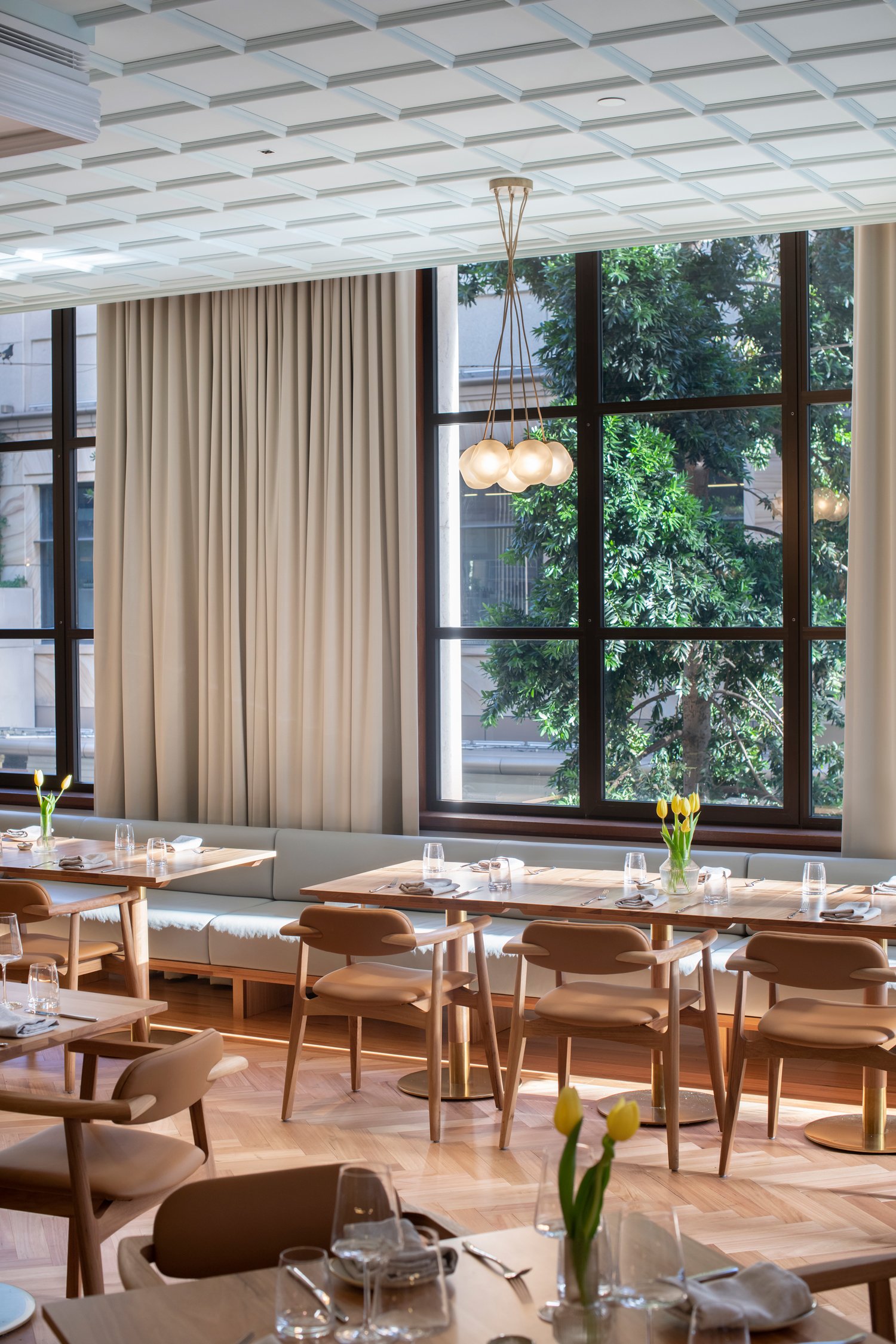Elegant dining area with large window views of trees, beige curtains, a modern chandelier with multiple globes, and tables set with water glasses, silverware, and small flower arrangements of yellow tulips.