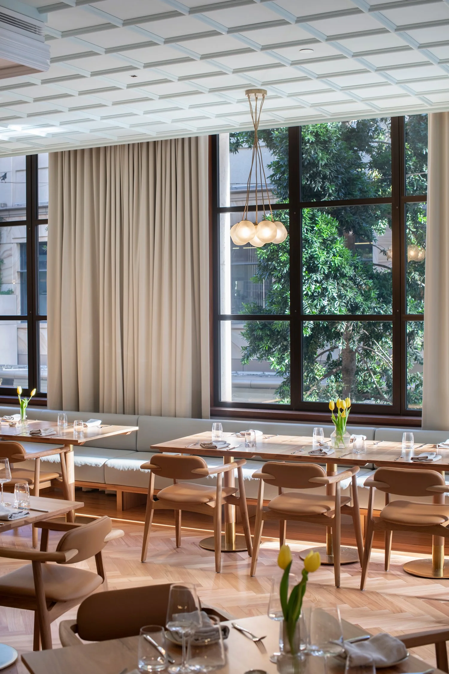 Dining area with large window views of trees, beige curtains, a modern chandelier with multiple globes, and tables set with water glasses, silverware, and small flower arrangements of yellow tulips.