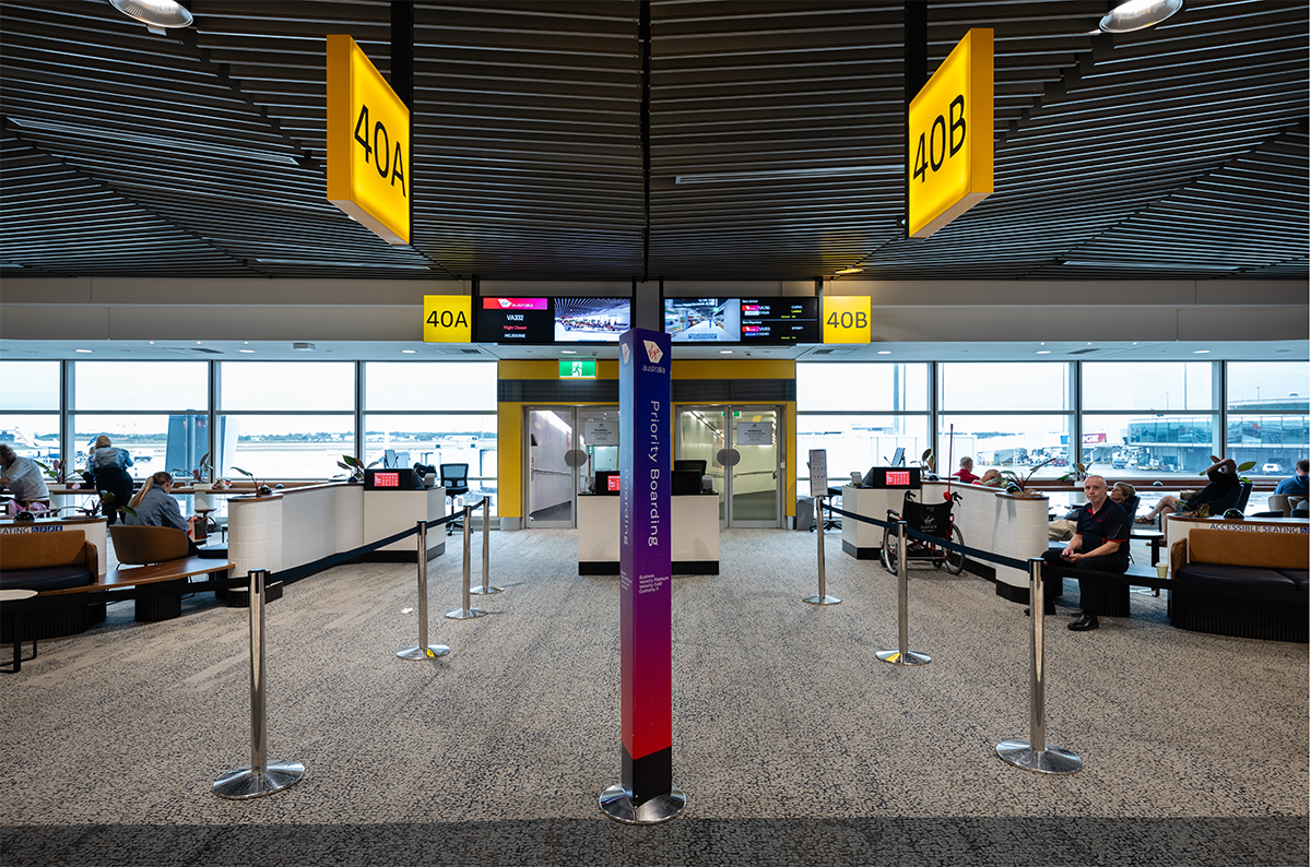 Airport gate area with yellow signs for gates 40A and 40B, seating areas, people waiting, and a view of the tarmac through large windows. Brisbane Airport Domestic Terminal. Retail Interior Design Australia