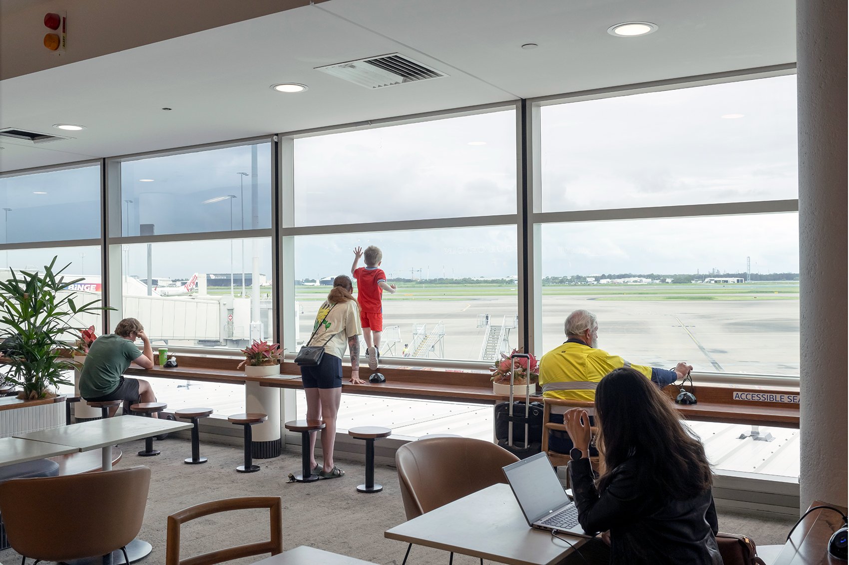 Interior of an airport lounge with people seated, looking out at the tarmac through large windows. A child climbs a windowsill, while others are engaged with laptops and phones. Brisbane Airport Domestic Terminal. Retail Interior Design Australia