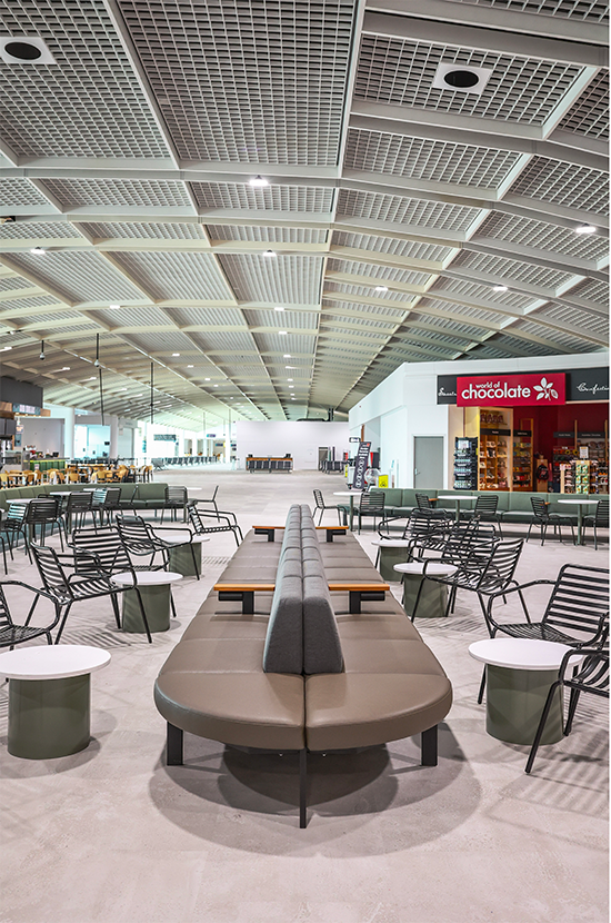 Empty airport lounge with modern chairs and a central curved bench, high ceiling with grid panel design, and a chocolate shop in the background.