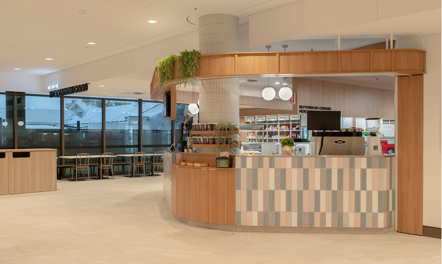 Prince Charles Hospital Foundation Common Good Cafe. Modern café in an indoor setting with bar-style seating, plants, and a wall of snacks and toiletries. Food and beverage Interior Design Brisbane Australia