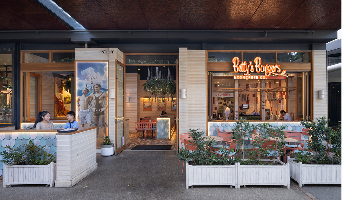Exterior view of Betty's Burgers & Concrete Co. restaurant with outdoor seating area, featuring a large neon sign, decorative plants, and customers interacting at tables.