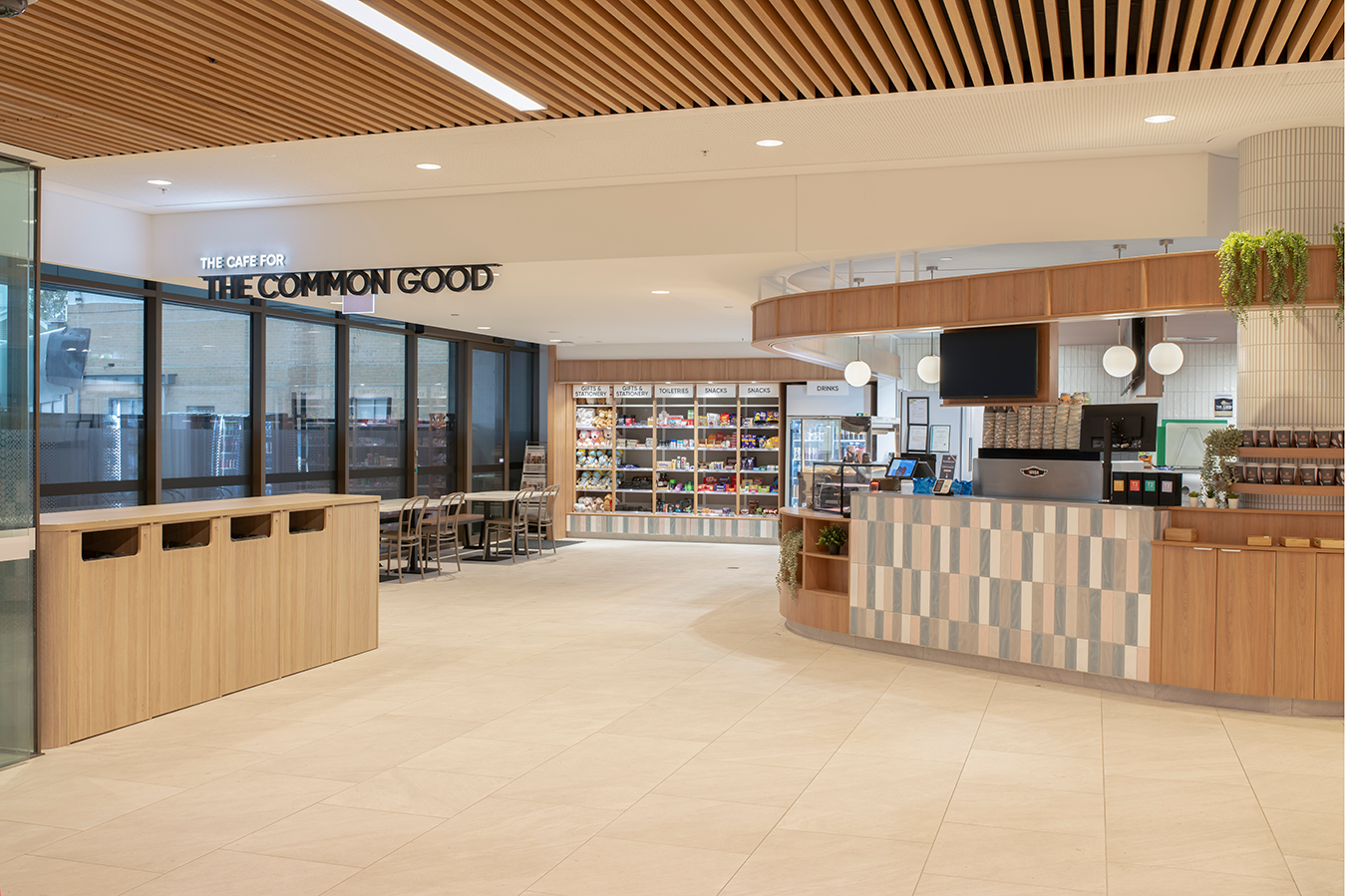 Prince Charles Hospital Foundation Common Good Cafe. Modern cafe interior with wood accents, shelves displaying snacks and drinks, and a counter area with a coffee machine. Food and beverage Interior Design Brisbane Australia