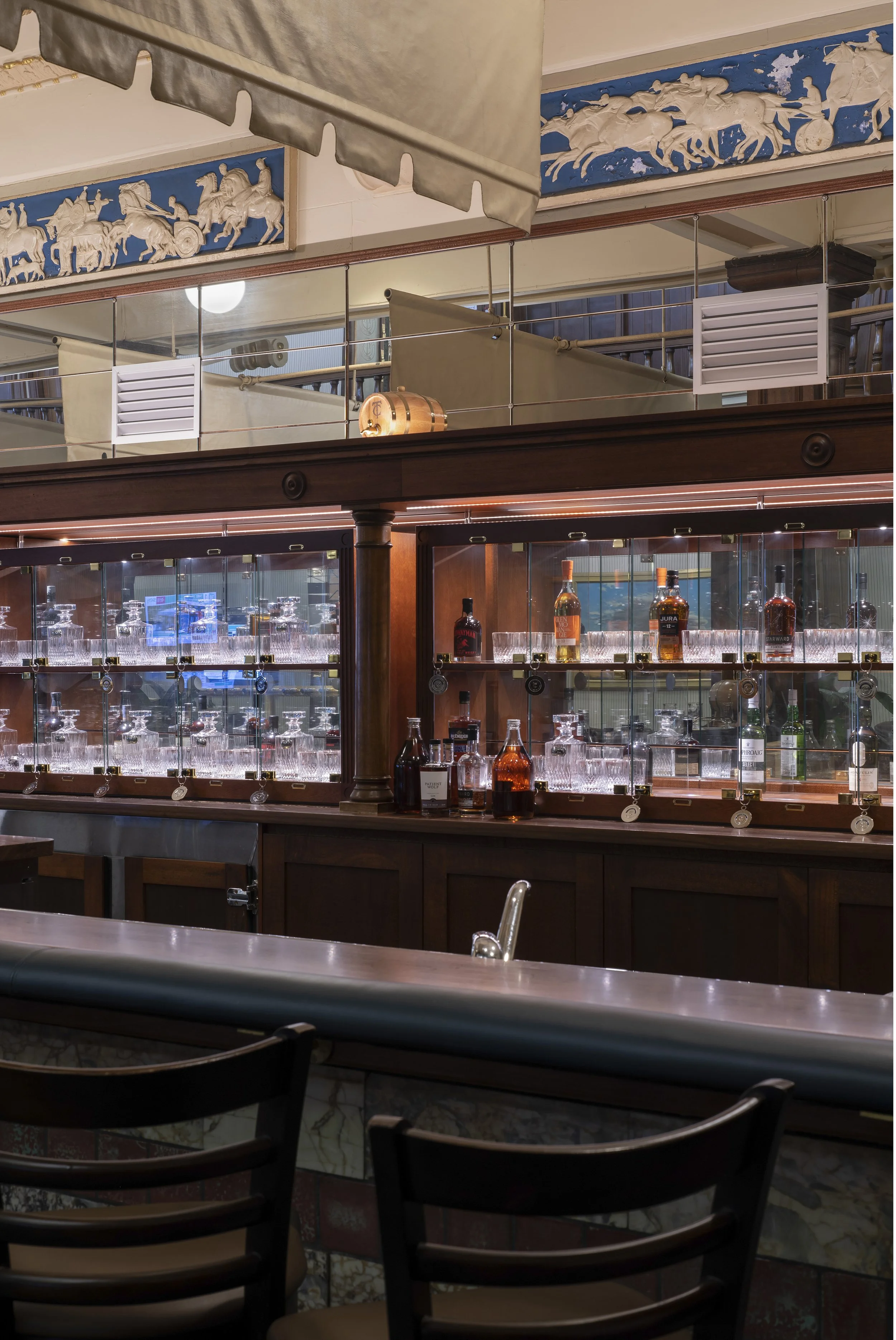 Interior of a bar with shelves displaying various liquor bottles and glasses, wooden cabinetry, and decorative frieze on the walls.