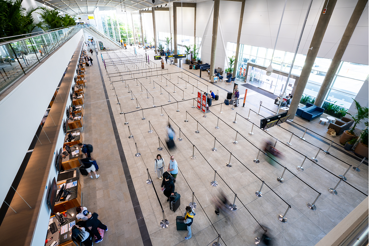 Cairns International T1 Airport. Indoor view of a modern airport terminal with check-in counters on the left, people lining up in roped queues, and large windows allowing natural light. Airport Interior Design Brisbane Australia