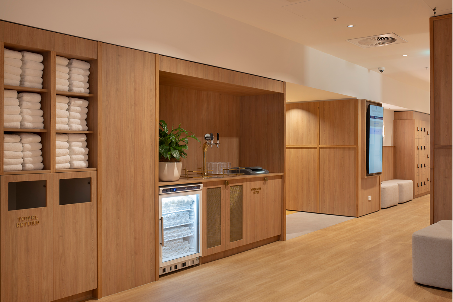 Tattersall's Club. A modern facility with wooden panels, stacked towels on shelves, a small fridge containing towels, a water dispenser, and a potted plant on a counter. Hospitality Interior Design Brisbane Australia