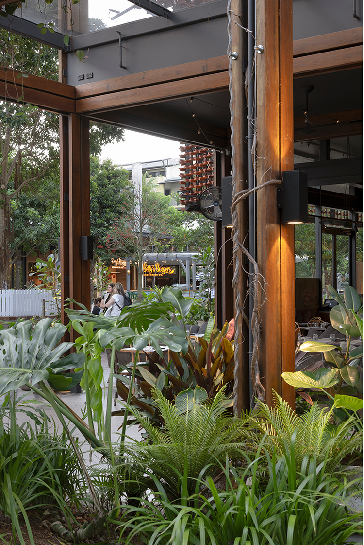 Outdoor dining area with wooden beams and lush green plants, featuring a glimpse of a burger restaurant sign in the background.