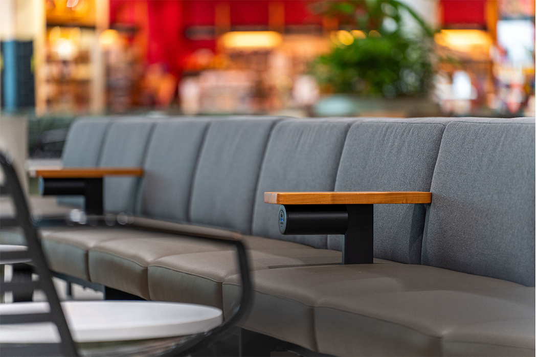Empty gray upholstered chairs with wooden armrests and built-in black speakers inside a modern public space, possibly an airport or lobby, with blurry background of colorful lights and shelves.