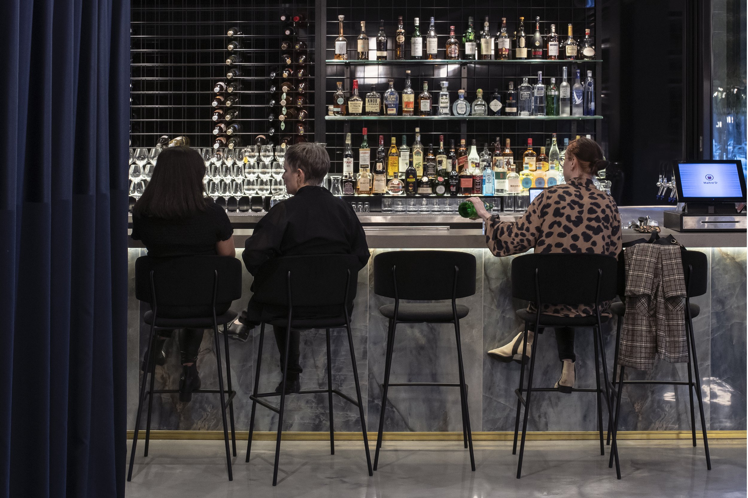 Bar interior with three patrons seated on high stools, a selection of liquor bottles on shelves behind the counter, and a digital device visible on the bar.