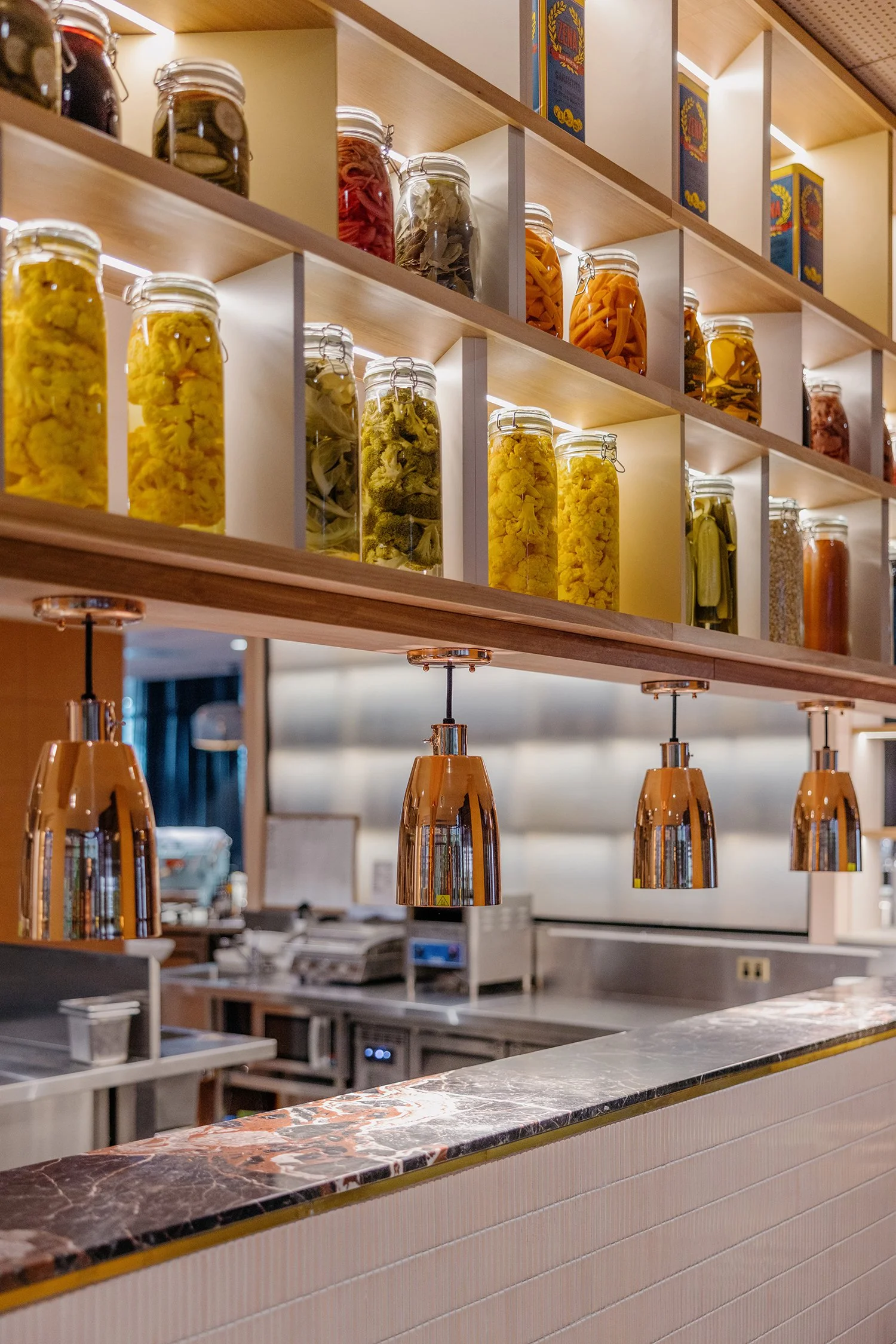 A bar counter with a marble top, copper pendant lights, and open shelves behind it filled with jars of pickled vegetables and boxed cereal.