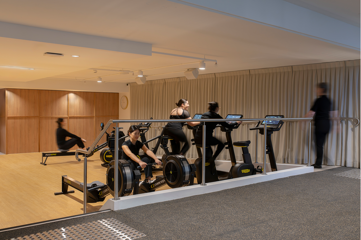A gym interior featuring people exercising on rowing machines and stationary bikes. The space has wooden flooring and a curtain backdrop. Tattersall's Club. Hospitality Hotel Restaurant Interior Design Brisbane Australia