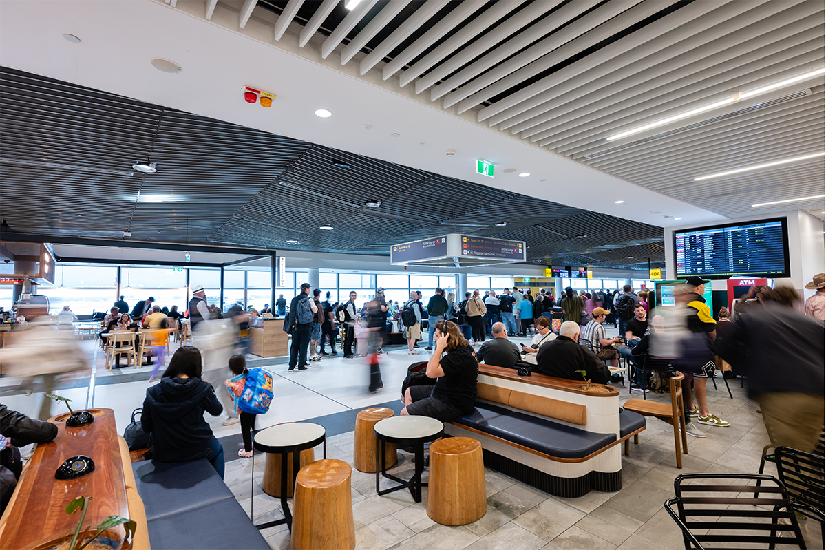 Busy airport terminal with travelers, seating areas, digital flight information display, and directional signs. Brisbane Airport Domestic Terminal. Retail Interior Design Australia