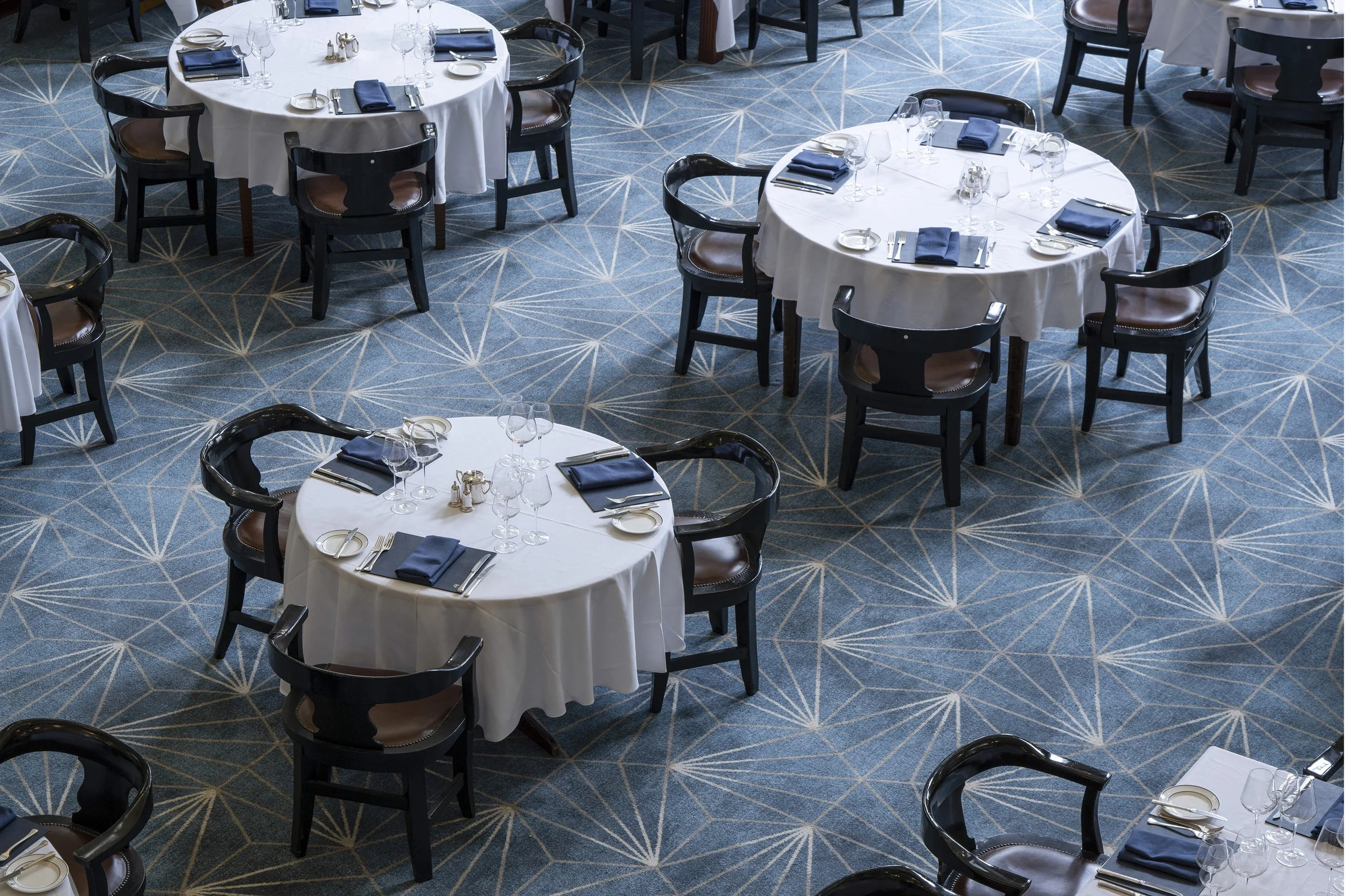 Elegant restaurant with round tables, white tablecloths, and blue napkins, set for dining with modern chairs on a patterned blue carpet.