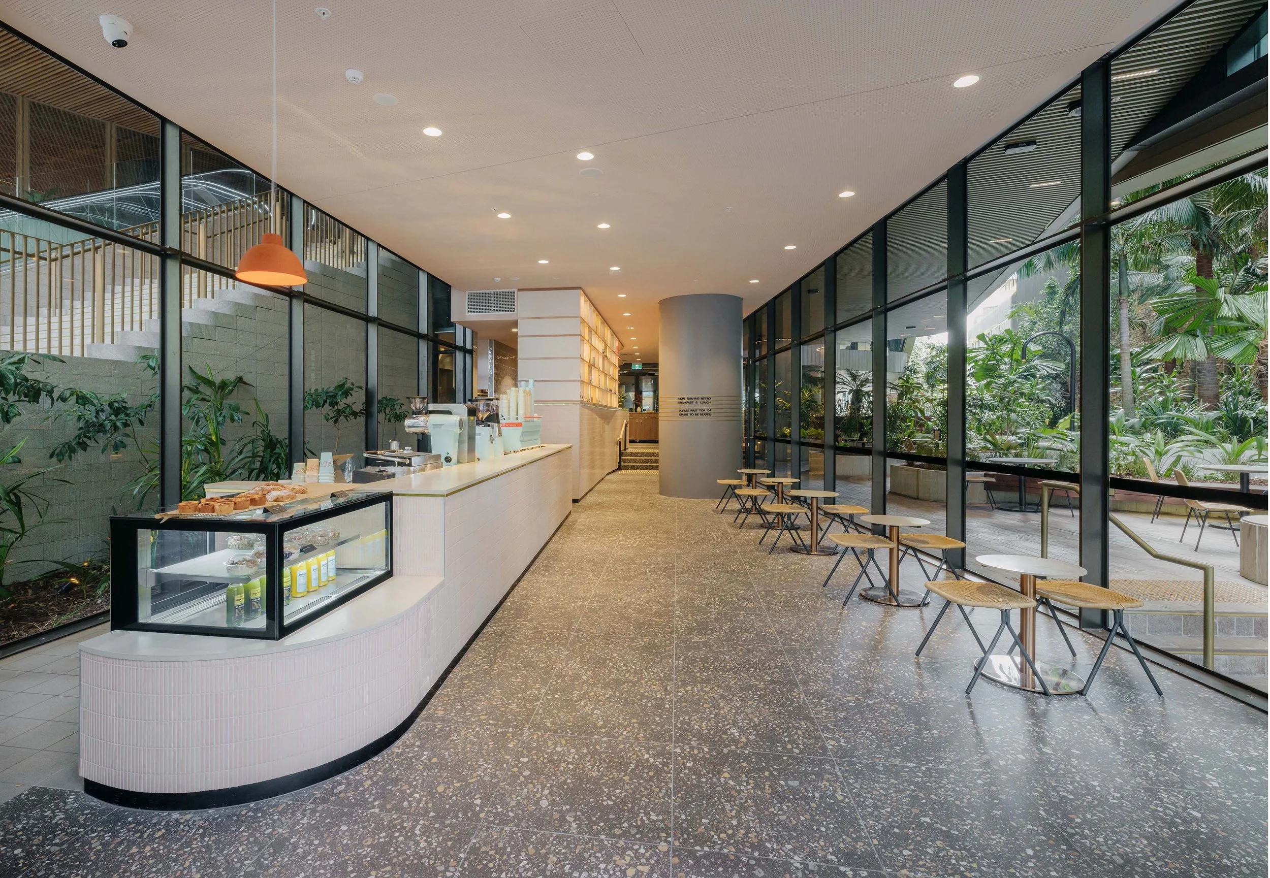 Modern cafe interior with a long counter, glass display case with pastries, seating area with small tables and stools, large windows, and indoor plants.