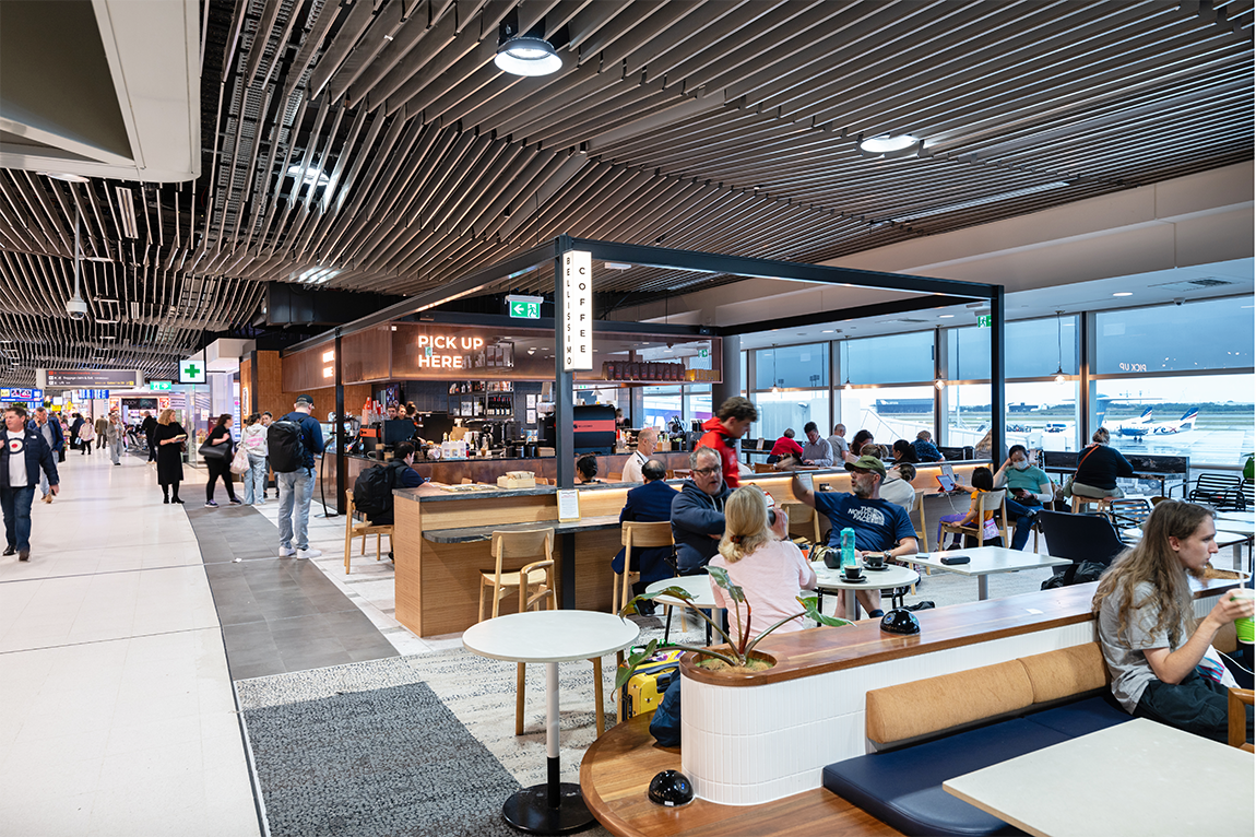 People in an airport cafe seating area, with travelers dining and a coffee pickup counter in the background. Brisbane Airport Domestic Terminal. Retail Interior Design Australia