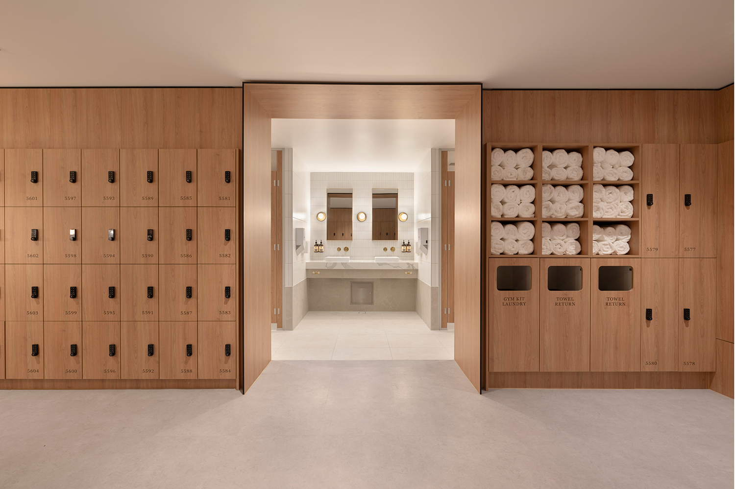 Modern locker room with wooden lockers, rolled white towels, towel return and laundry bins, and a view into a restroom with mirrors and sinks.