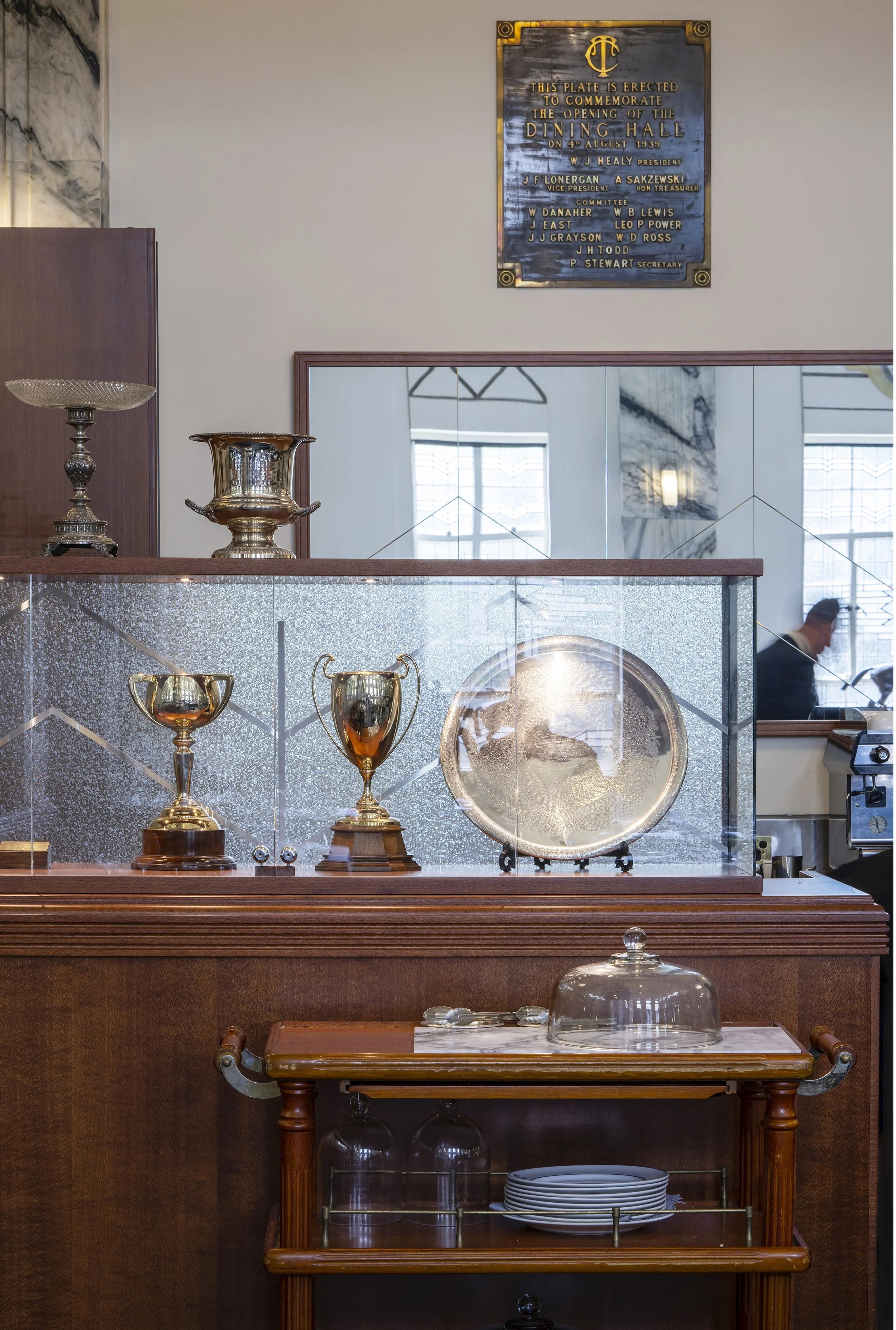 Display of trophies and a plaque on a wooden cabinet, with a glass-covered dish on a cart below and plates and glasses stored underneath.