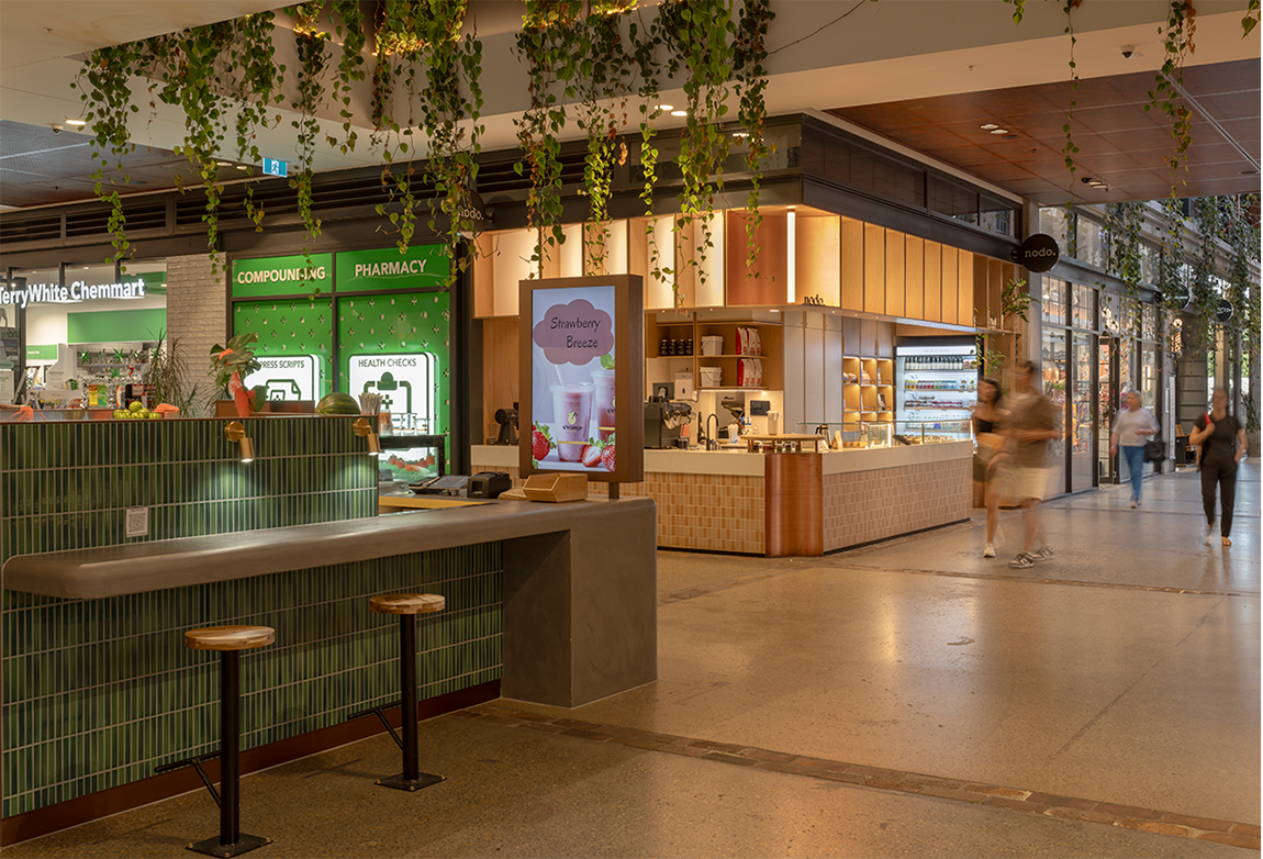Retail interior with pharmacy and cafe, hanging plants, and people walking.