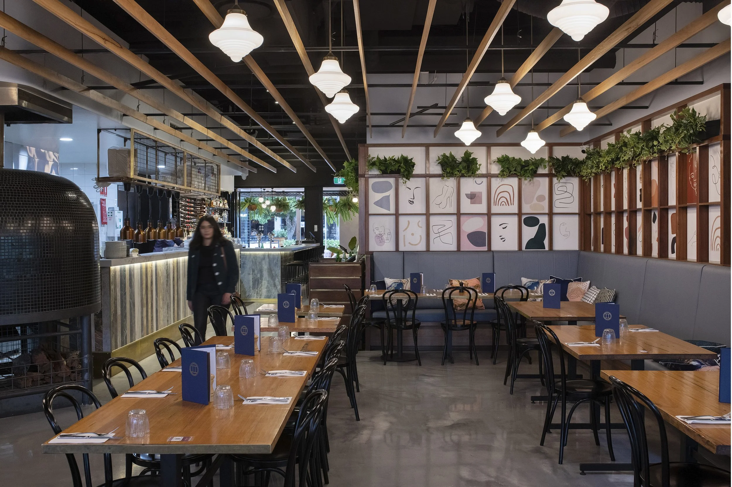 Interior of a modern restaurant with wooden tables, black chairs, and contemporary decor. Decorative plants and abstract art adorn the walls. Soft lighting and a welcoming atmosphere.