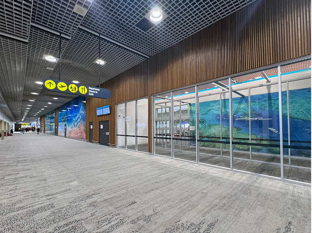 Airport corridor with signage for departures, check-in, and café; glass wall with ocean-themed mural; overhead lights and ceiling grid.
Townsville Airport. Interior Design Australia