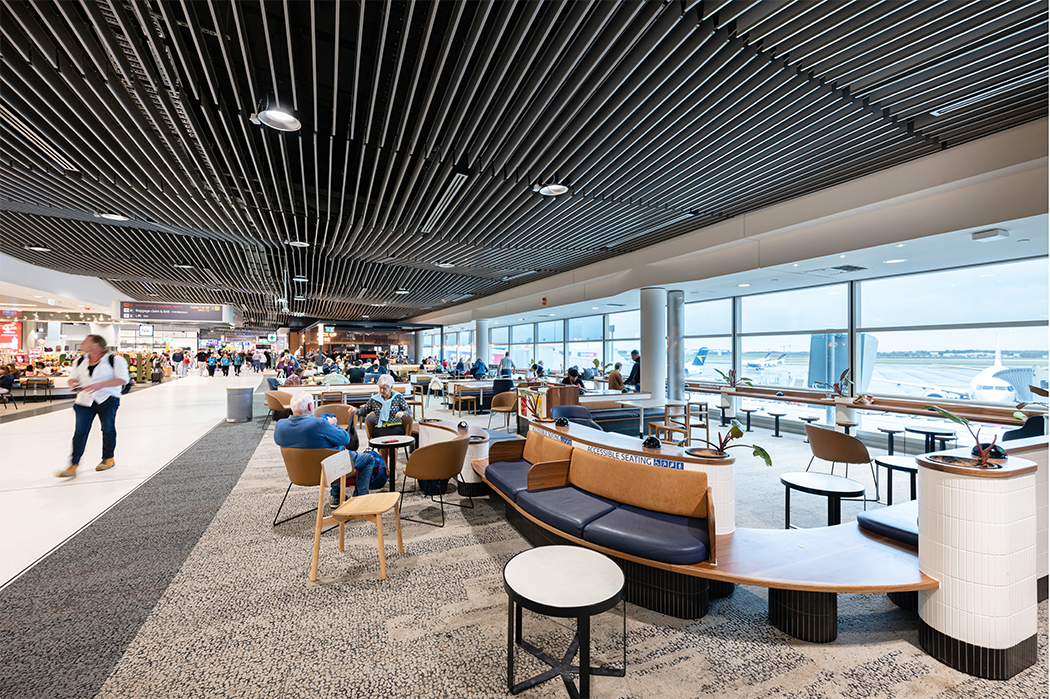 Modern airport terminal with passengers, seating areas, and large windows overlooking airplanes on the tarmac. Brisbane Airport Domestic Terminal. Retail Interior Design Australia