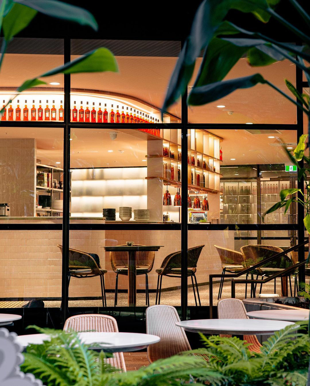 View of a restaurant or cafe interior seen through large window with plants in foreground; inside, shelves with bottles, a counter with plates, and tables and chairs for dining.