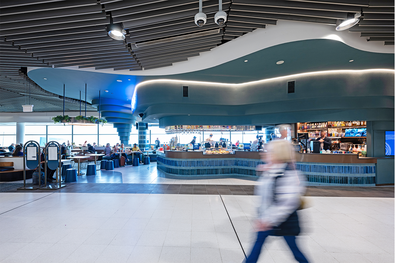 Airport lounge with modern design, featuring a bar and seating area, with people sitting and walking, blurred motion. Brisbane Airport Domestic Terminal. Retail Interior Design Australia