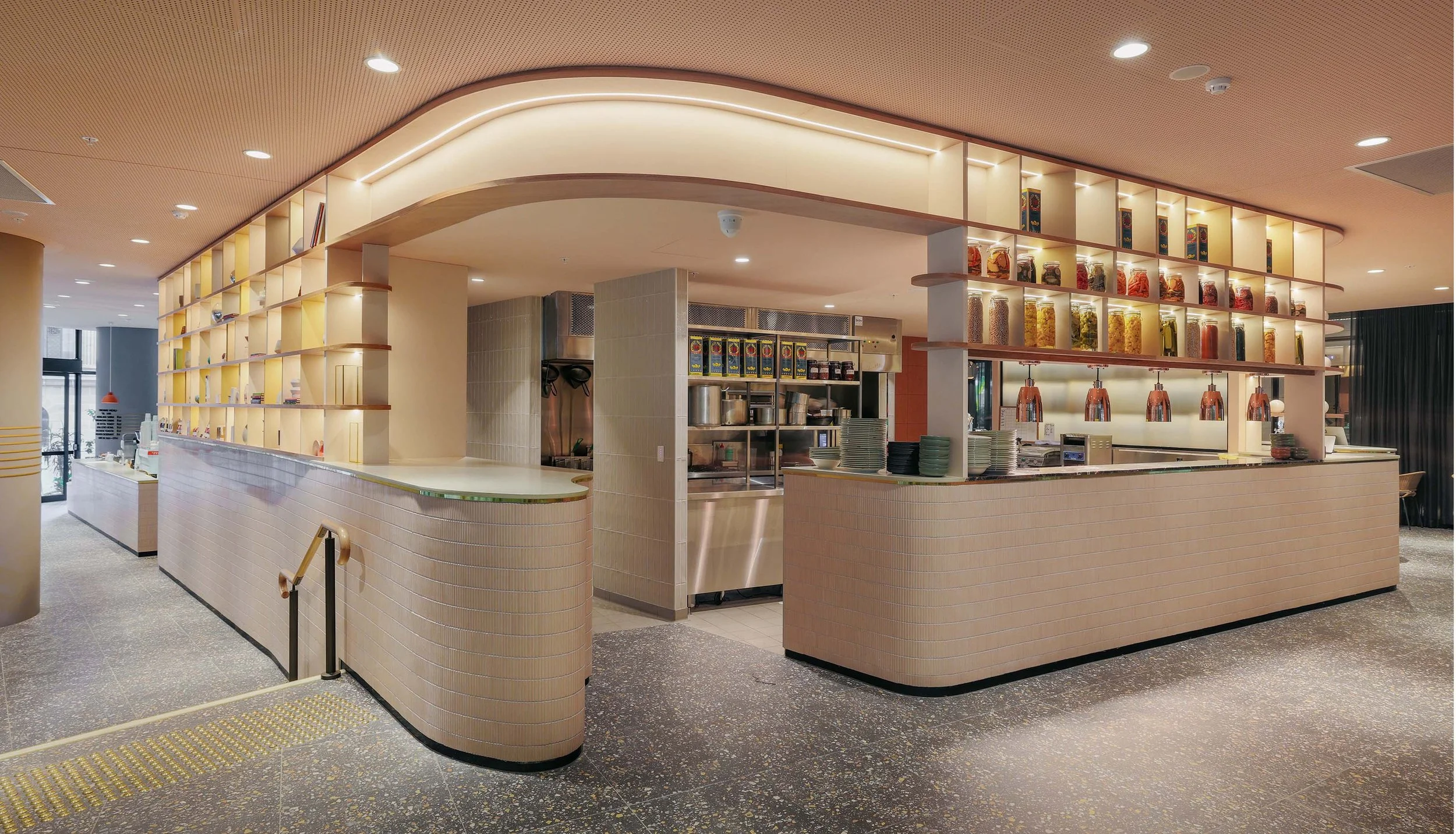 Modern restaurant interior with open kitchen and countertop, surrounded by shelves with jars and decorative items.