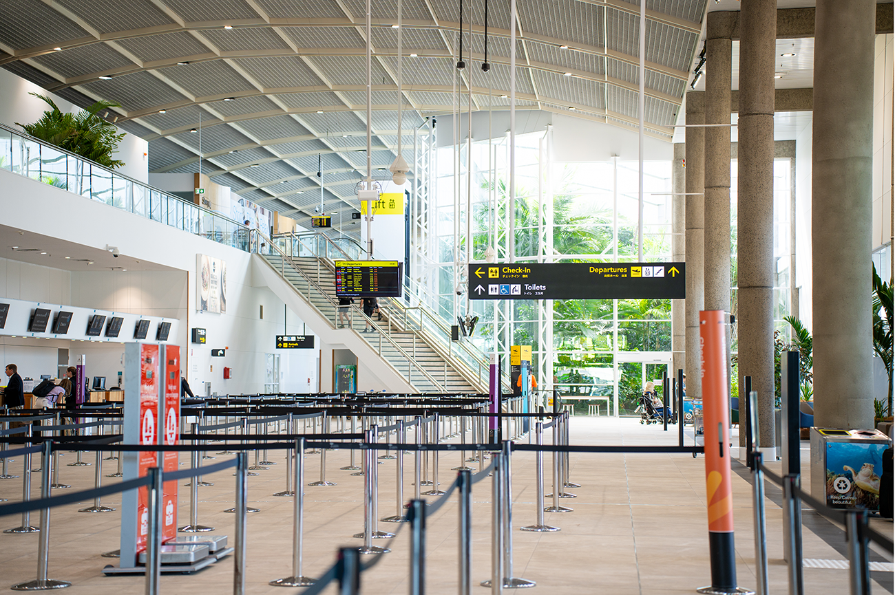 Cairns International T1 Airport. Interior of an airport terminal with check-in counters, a departures sign, and people standing in a queue; large windows with greenery outside. Airport Interior Design Brisbane Australia