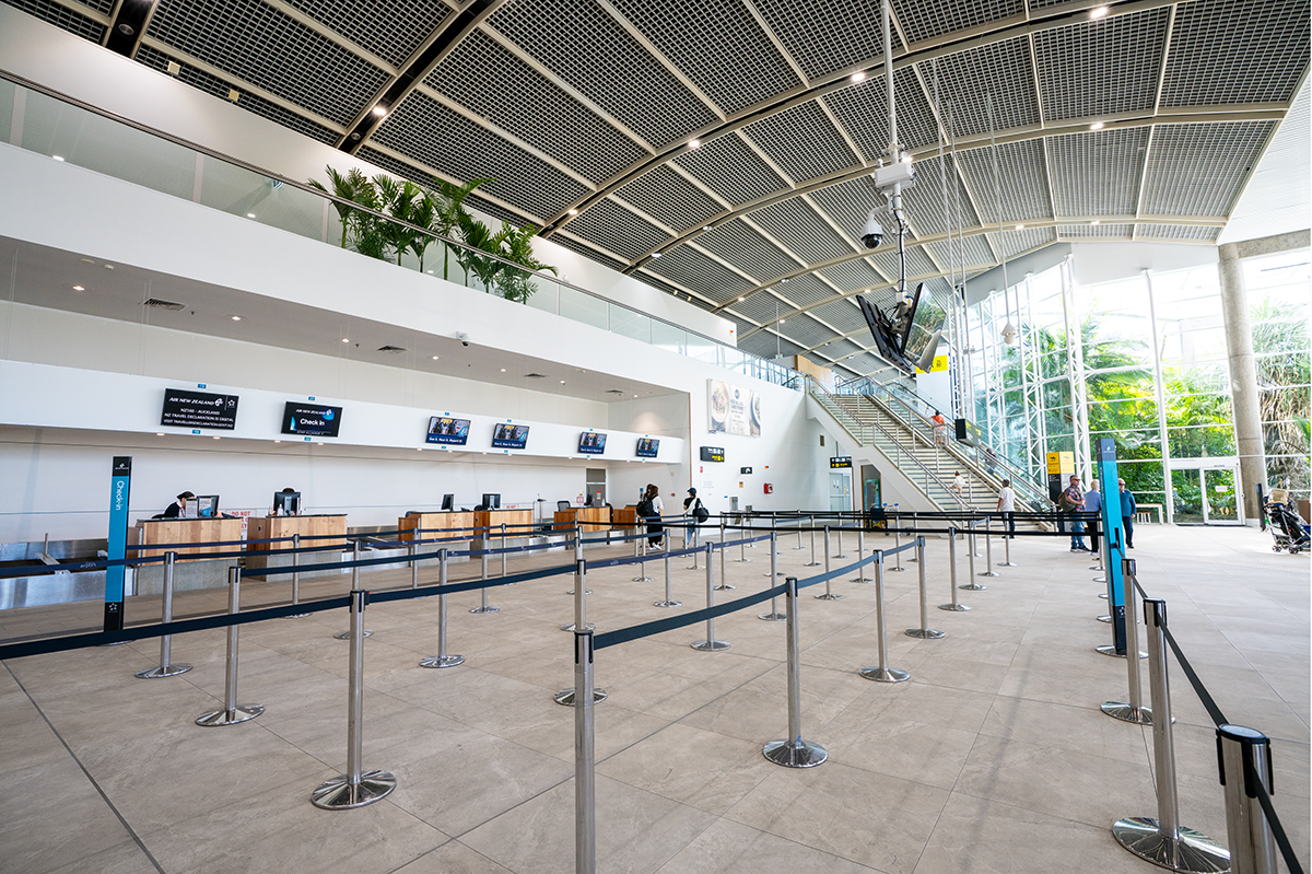 An empty airport check-in area with multiple counters and digital screens. There are queue barriers forming lines, a staircase in the background, and a glass wall with natural light. Few people are present in the distance. Hospitality Interior Design