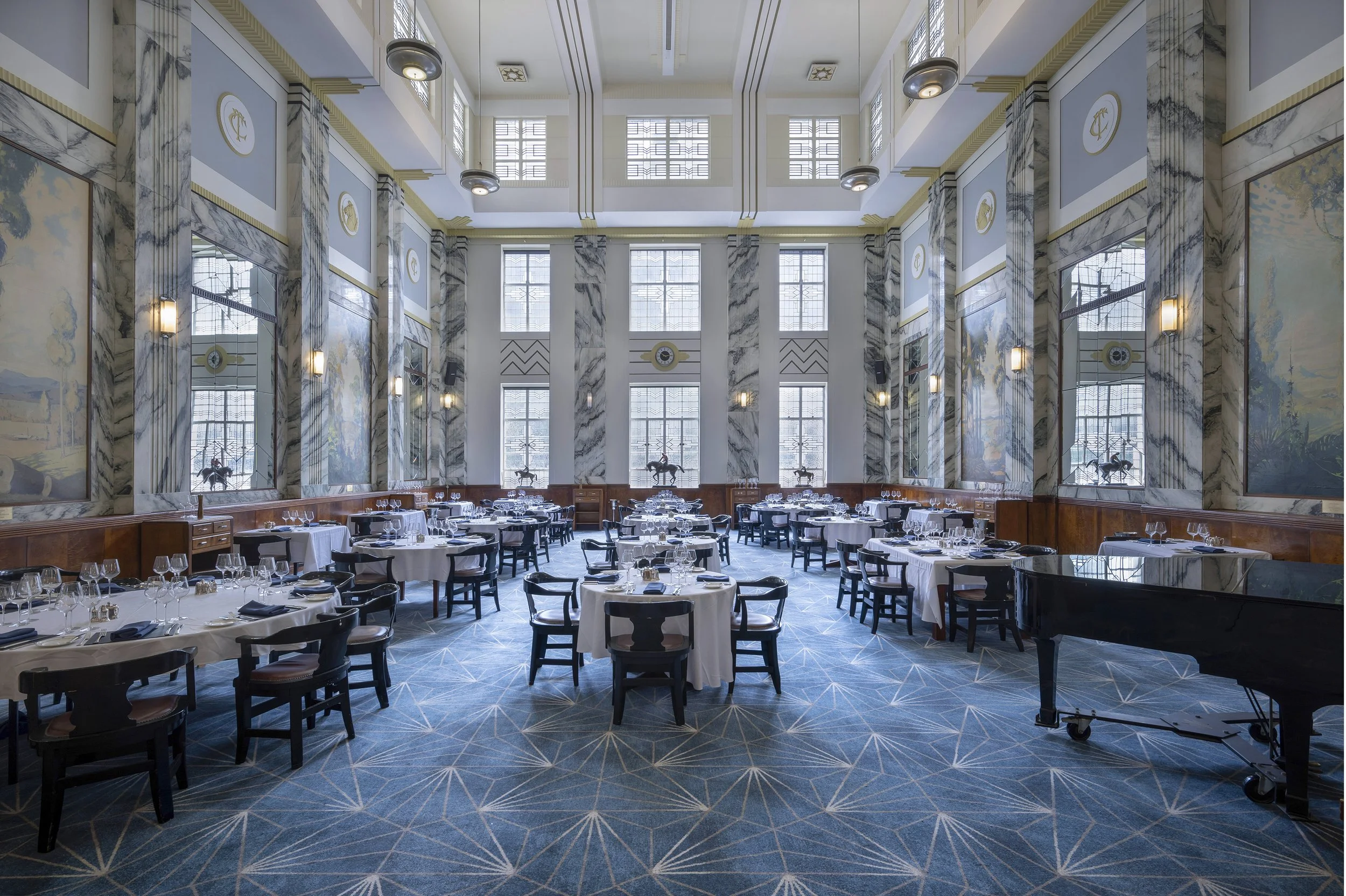 Elegant dining room with high ceilings, large windows, blue patterned carpet, round tables with white tablecloths, black chairs, and a grand piano.